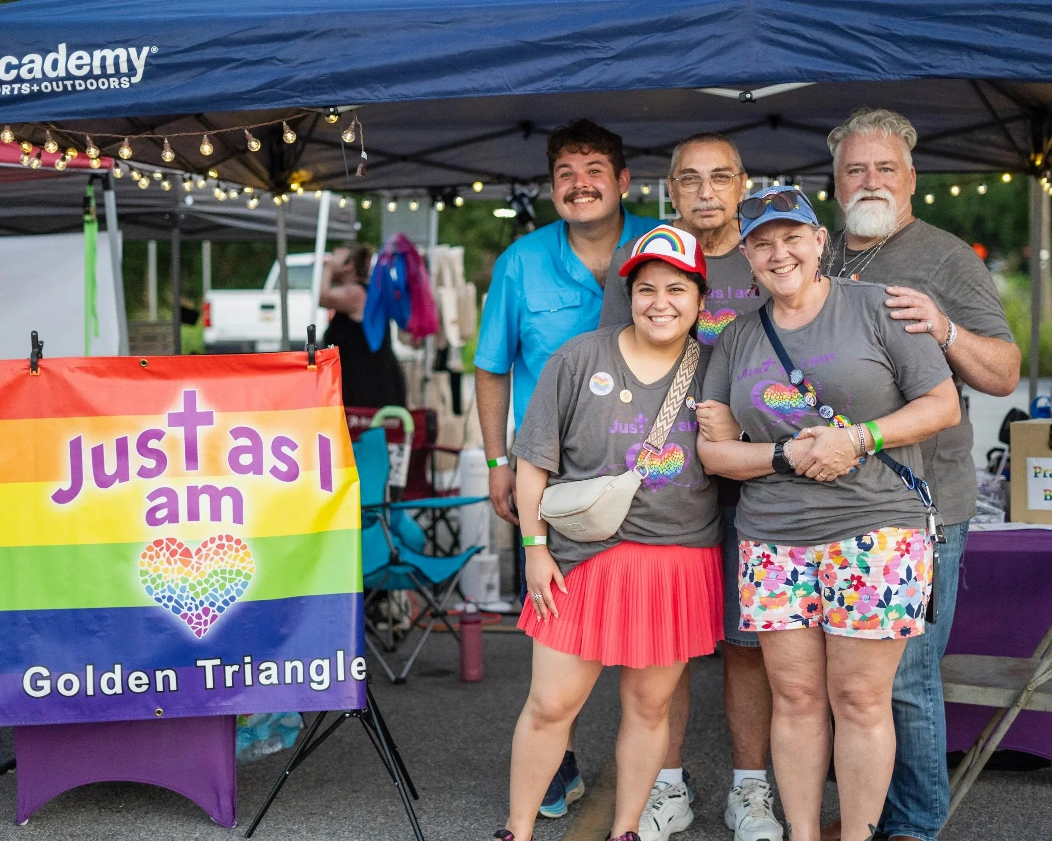 People standing under a canopy at an outdoor event, smiling. There are rainbow-colored LGBTQ+ pride buttons and a rainbow hat. A rainbow heart-shaped logo is visible on a sign that reads 'Just as I am'.