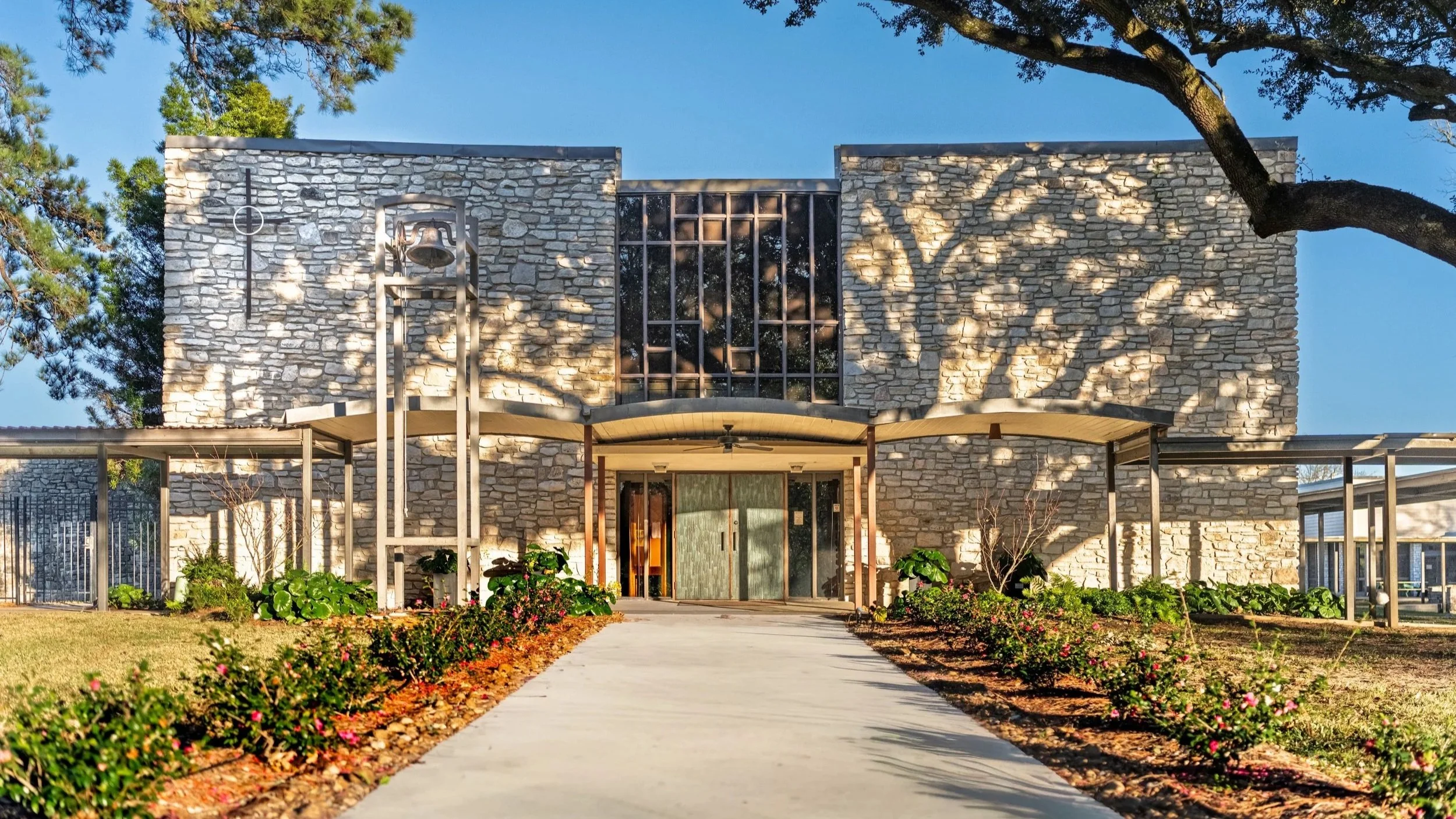 A wide cement walkway leading to the double doors of a sandstone building with double doors on the front.  The walkway is lined with shrubbery. There is a bell tower on the left side of the photo.