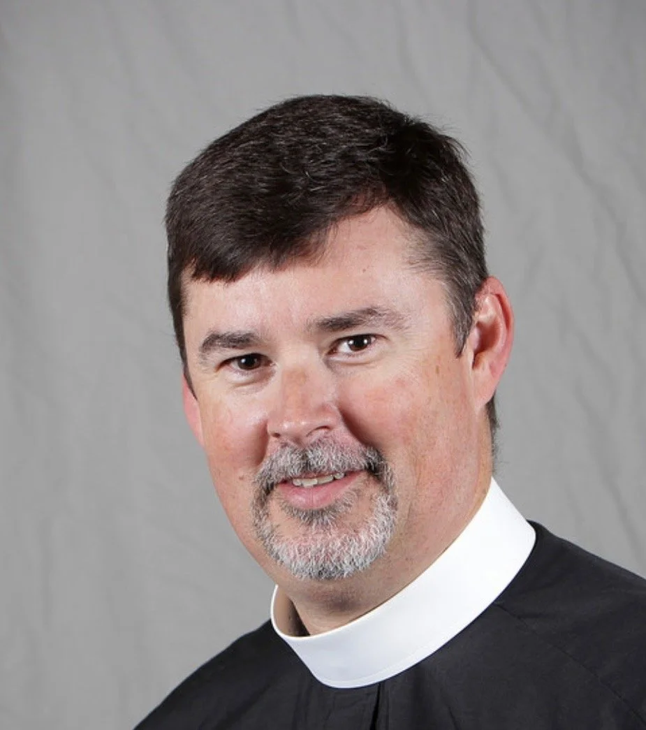 Head shot of a man with dark hair parted on one side with a graying goatee wearing a black shirt with a priest collar.
