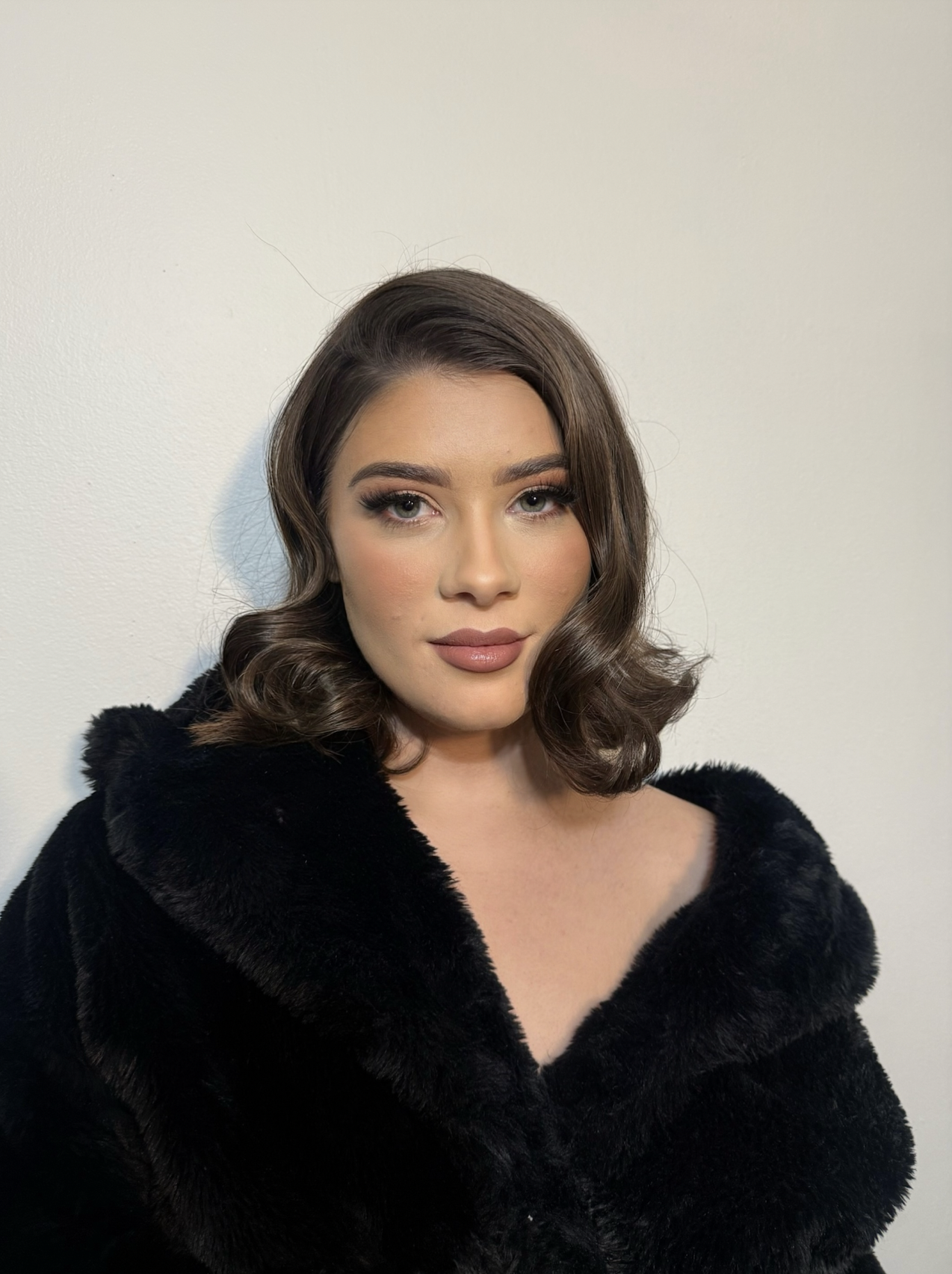 A young woman with dark brown, shoulder-length curled hair, wearing makeup with neutral tones, and a black fur coat, standing against a plain white wall.