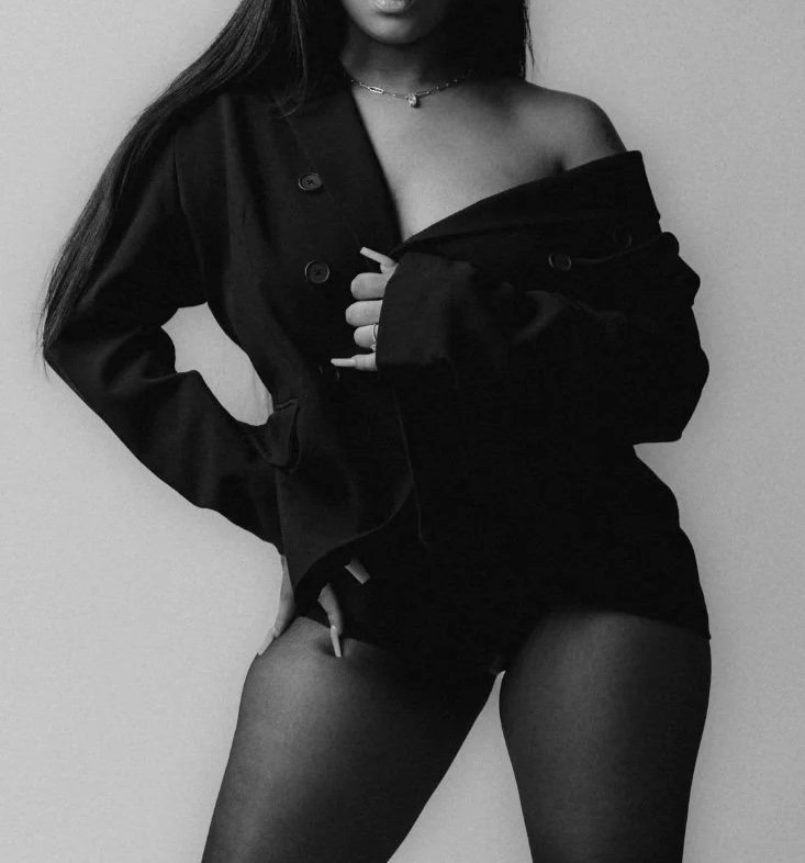 A woman in black clothing poses against a plain wall, with one hand on her hip and the other adjusting her coat, wearing sheer tights and a necklace.