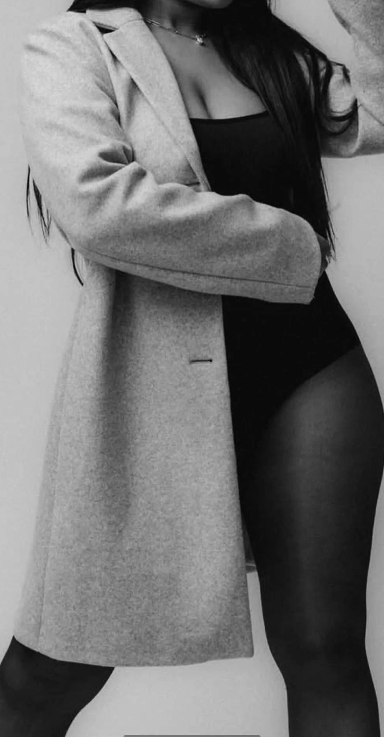 A woman wearing a long coat, a black top, and black leggings, with long dark hair partially covering her face in a black and white photo.