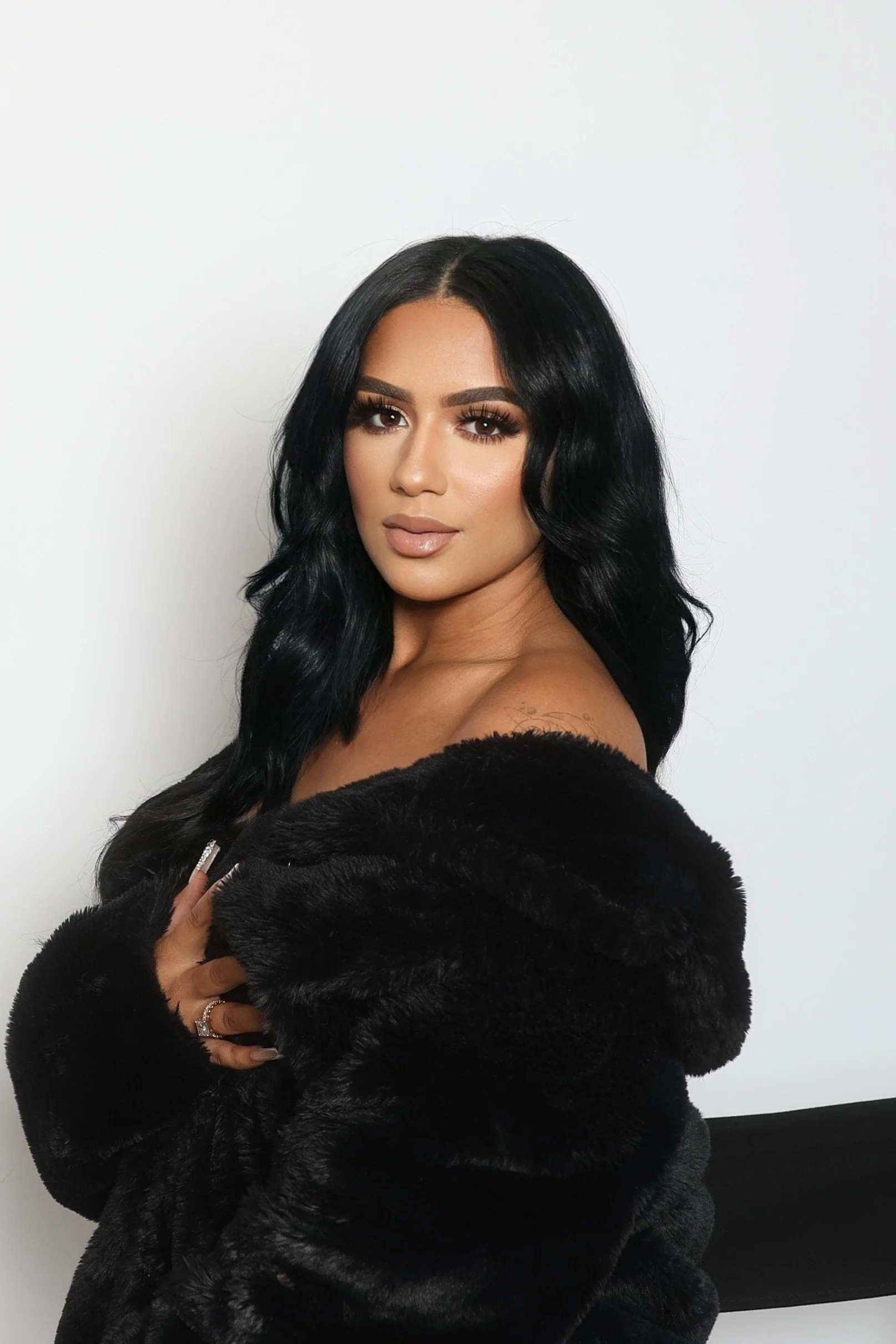 A woman with long black wavy hair, makeup, and nude lipstick, wearing a black fur coat, looking at the camera against a white background.