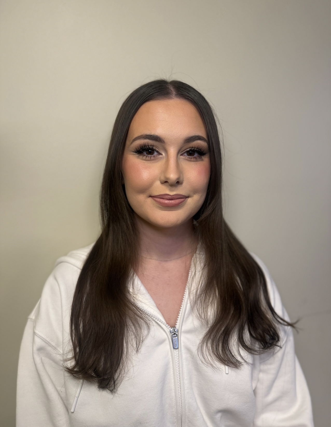 Portrait of a young woman with long brown hair wearing a white zip-up hoodie, smiling softly against a plain light-colored background.