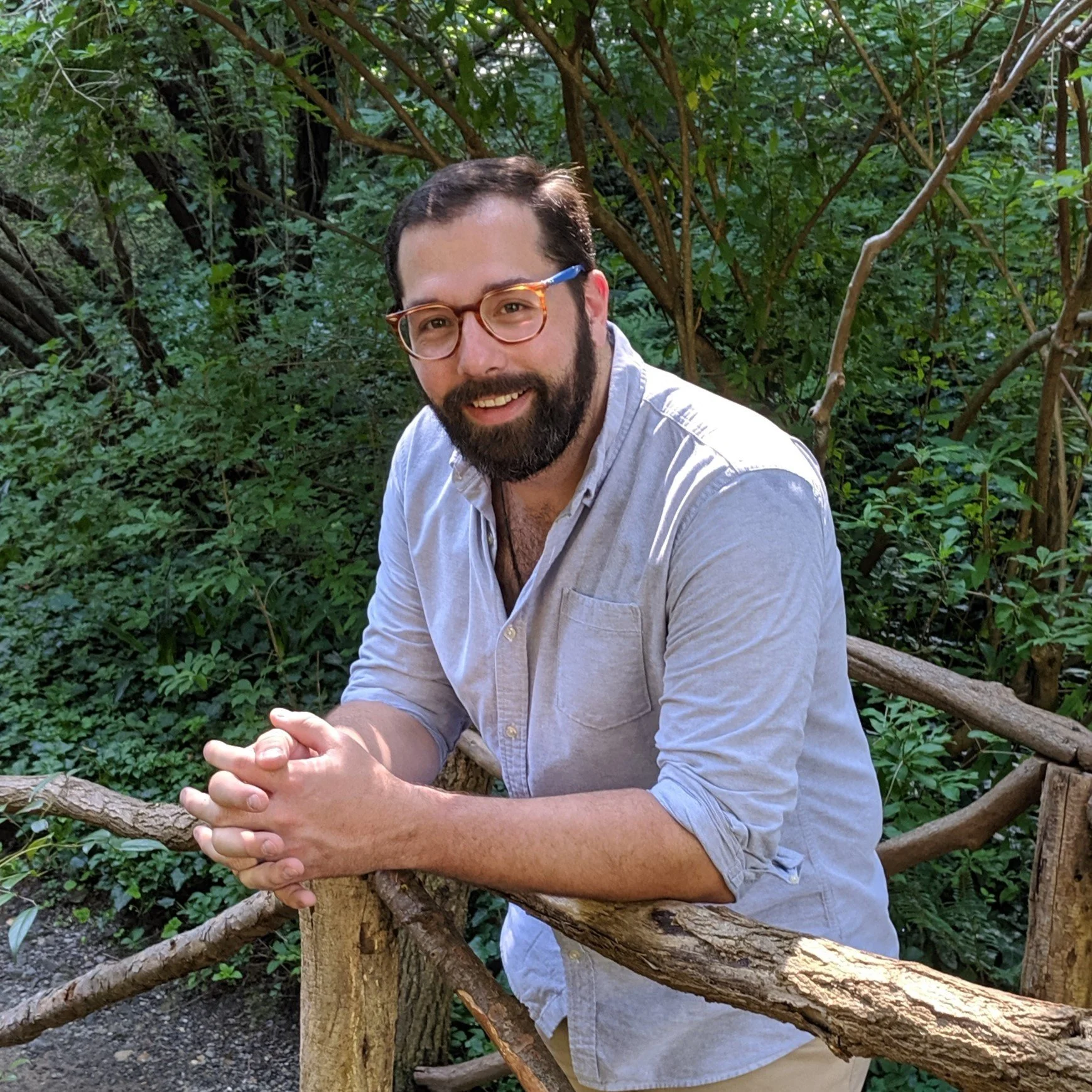 A man with glasses and a beard, wearing a light gray button-up shirt, is outdoors on a wooden bridge in a green, leafy forest. He is smiling and leaning on the railing with his hands clasped.
