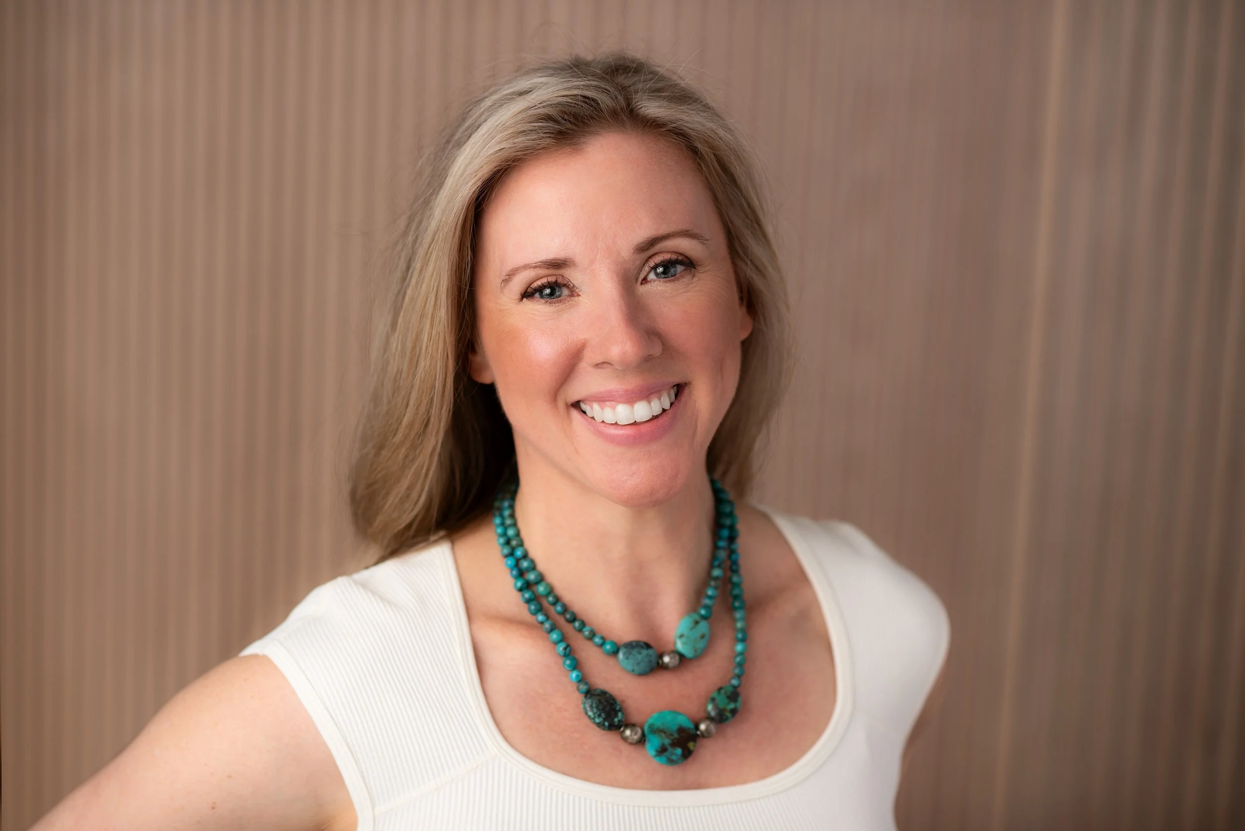 A smiling woman with blonde hair wearing a white top and turquoise beaded necklace standing in front of a wooden background.