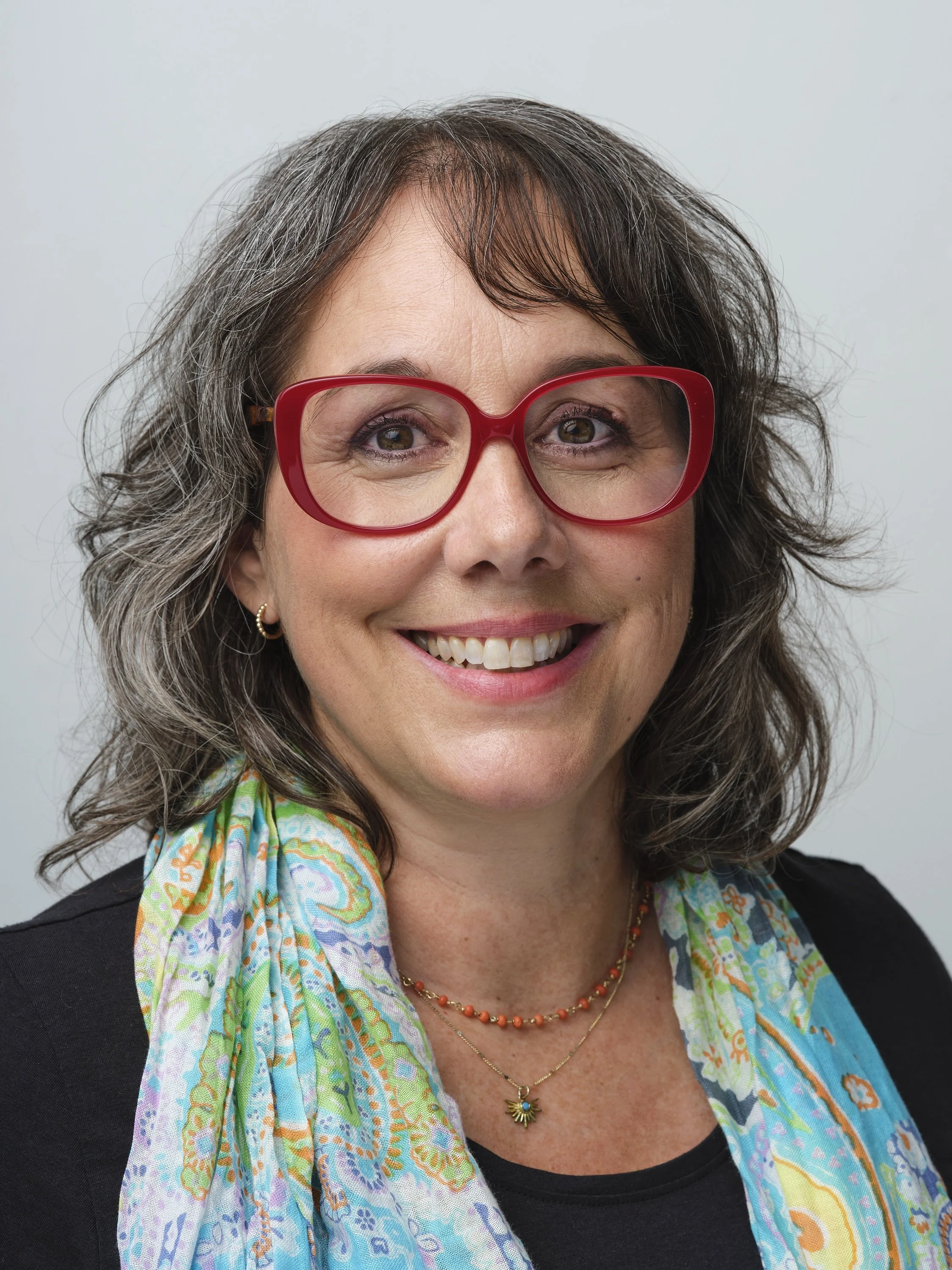 A woman with curly brown hair, wearing red glasses, smiling, with colorful scarf, layered necklaces, and earrings, against a light gray background.