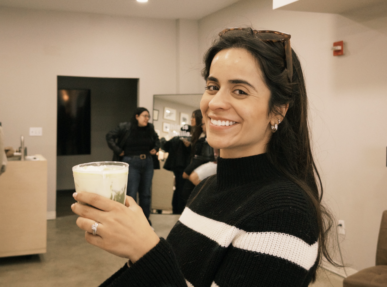 A woman smiling and holding a glass of a green and white beverage in an indoor setting with other people in the background.