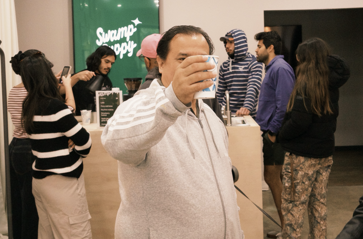 A group of people standing in line at a coffee shop, with one man in the foreground holding a cup and smiling.