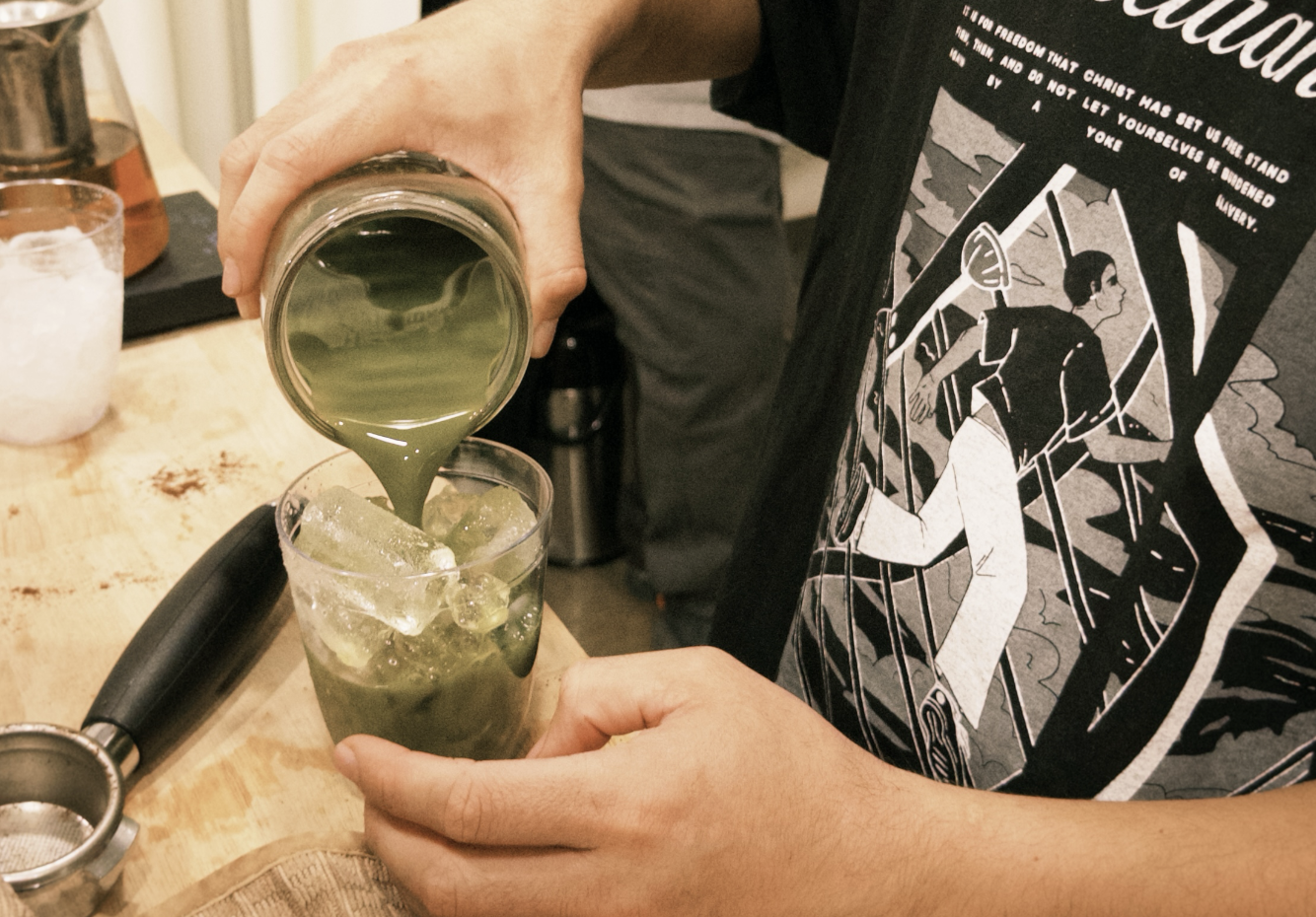 Person pouring matcha green liquid into a glass with ice on a wooden bar counter.