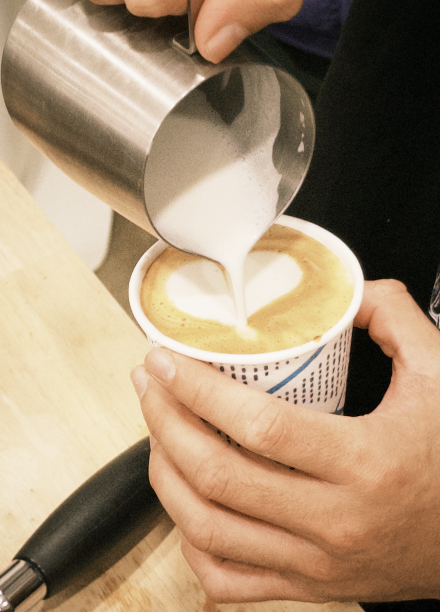 Barista pouring steamed milk into a cup of coffee, creating latte art with foam.