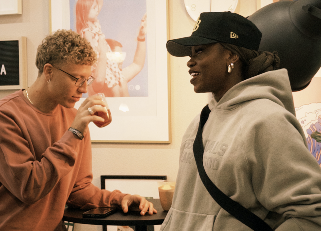 Two women conversing at a cafe or art gallery, one holding a drink, with colorful artwork on the wall behind them.
