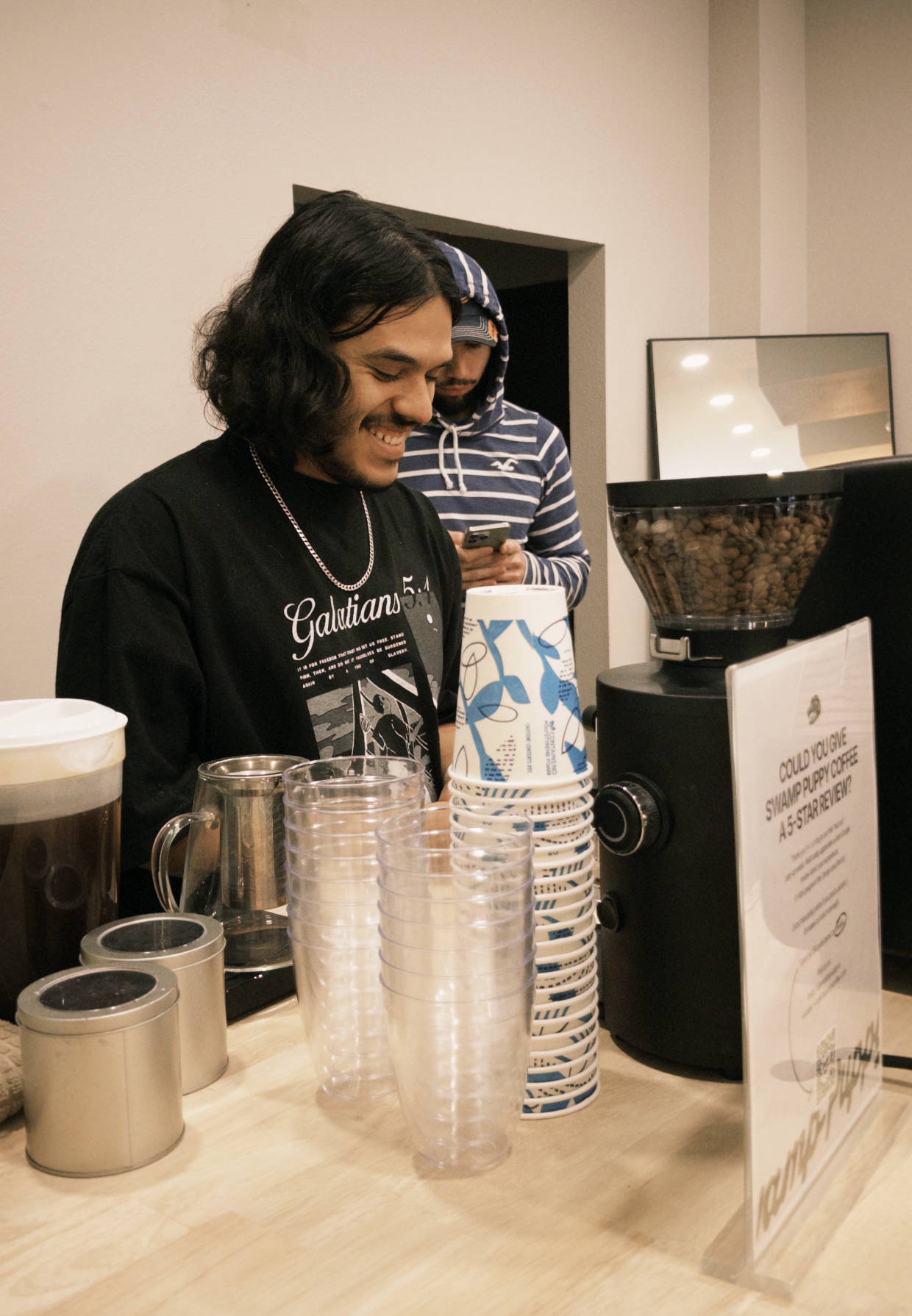 Two young men standing behind a coffee counter, one smiling and looking down, the other looking at a phone, with coffee brewing equipment and cups on the counter.
