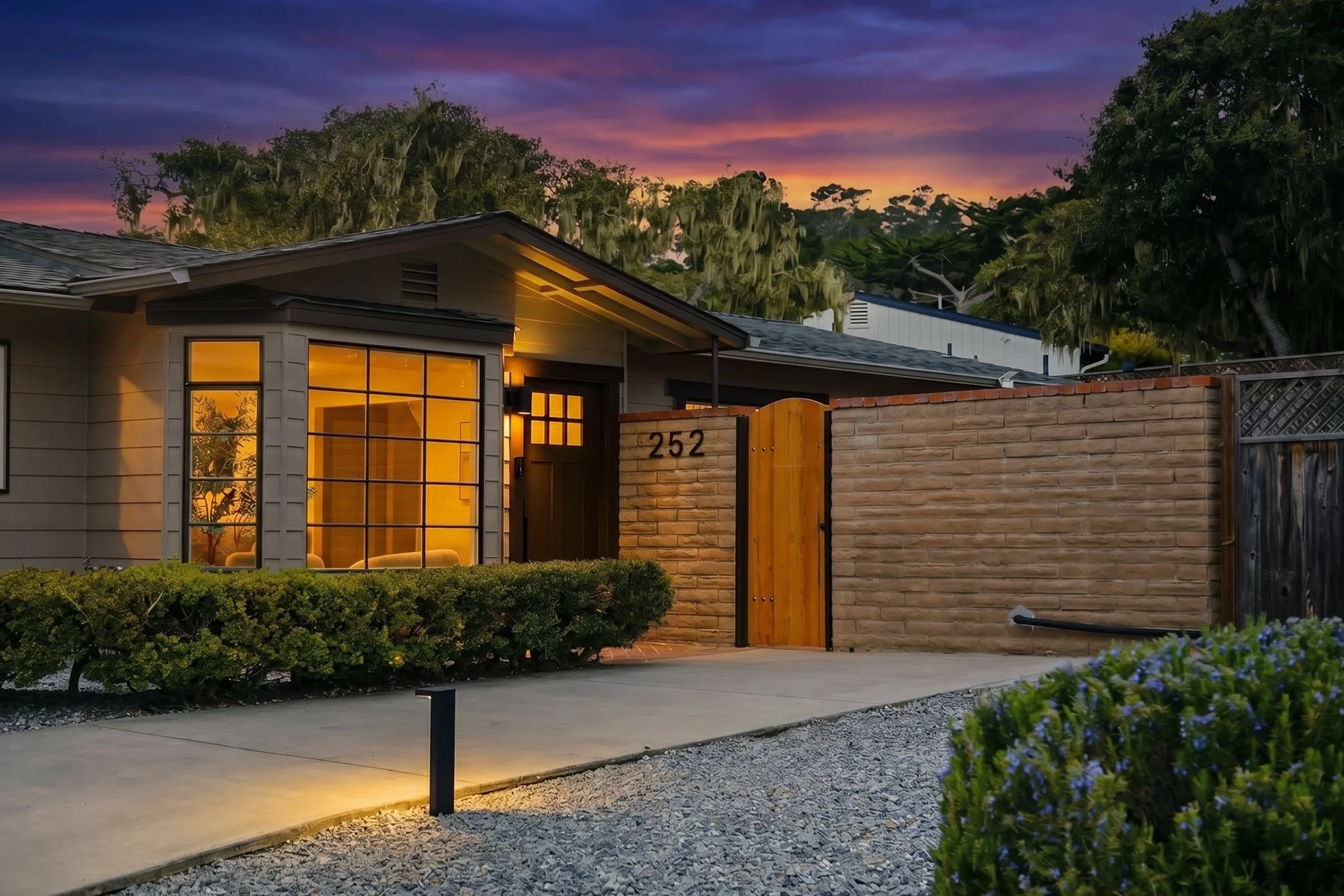 Modern house with illuminated interior, brick wall, and garden, during sunset with colorful sky and trees in the background.