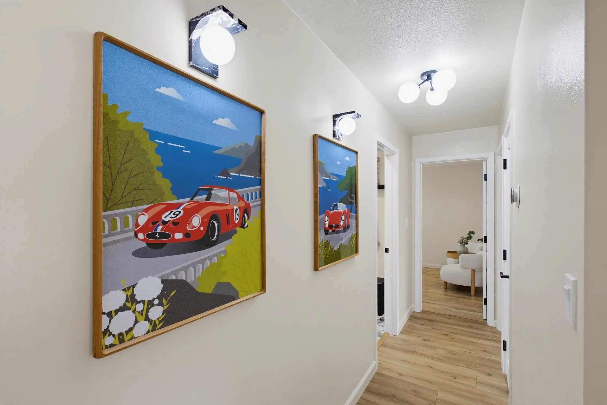 Hallway with framed car race-themed artwork on the wall, modern light fixtures, white walls, and wooden flooring leading into a living room area with a white armchair and a side table with a plant.
