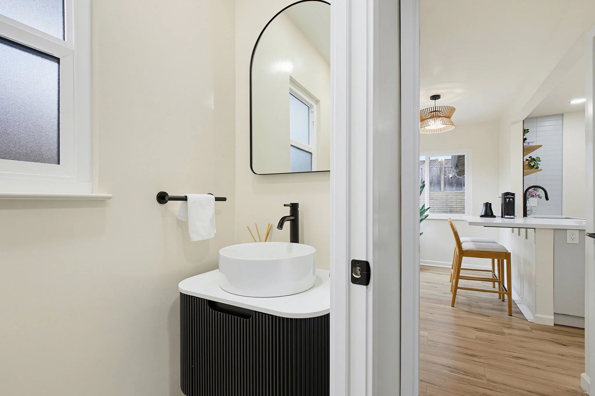 A modern bathroom with a round vessel sink, black faucet, and black vanity. A large mirror is mounted on the wall, and a window with frosted glass. Part of the adjacent kitchen with wooden chairs, white countertops, and a pendant light fixture are visible.
