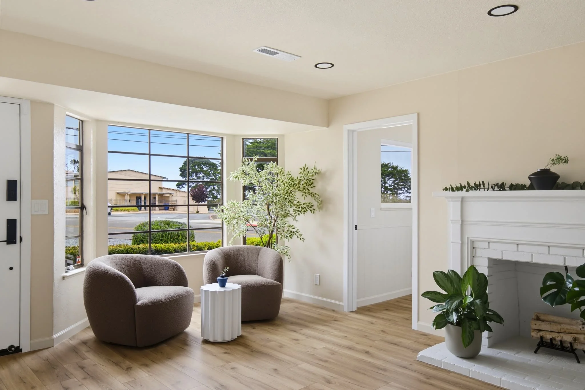 Interior of a living room with two brown armchairs, a white side table, large windows, a fireplace decorated with plants, and hardwood floors.