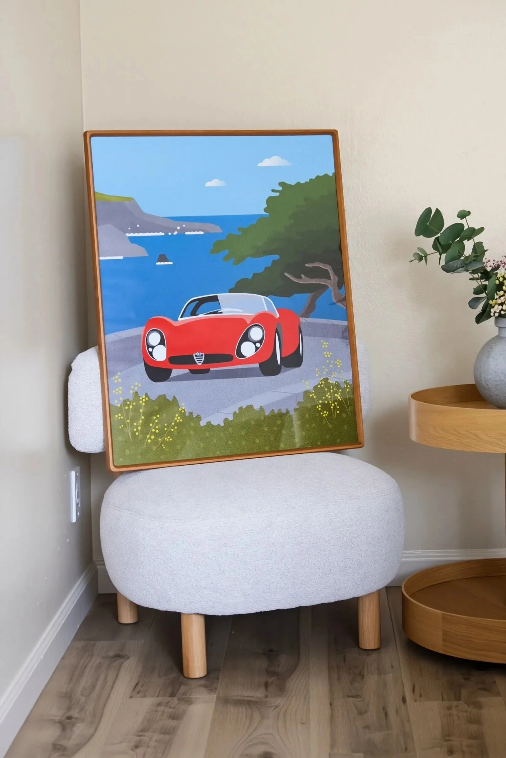 A framed colorful landscape painting of a coastal scene with a red sports car in the foreground, displayed on a white cushioned chair with wooden legs in a room with light-colored walls and wooden flooring. Next to the chair is a small wooden side table with a potted plant with green leaves.