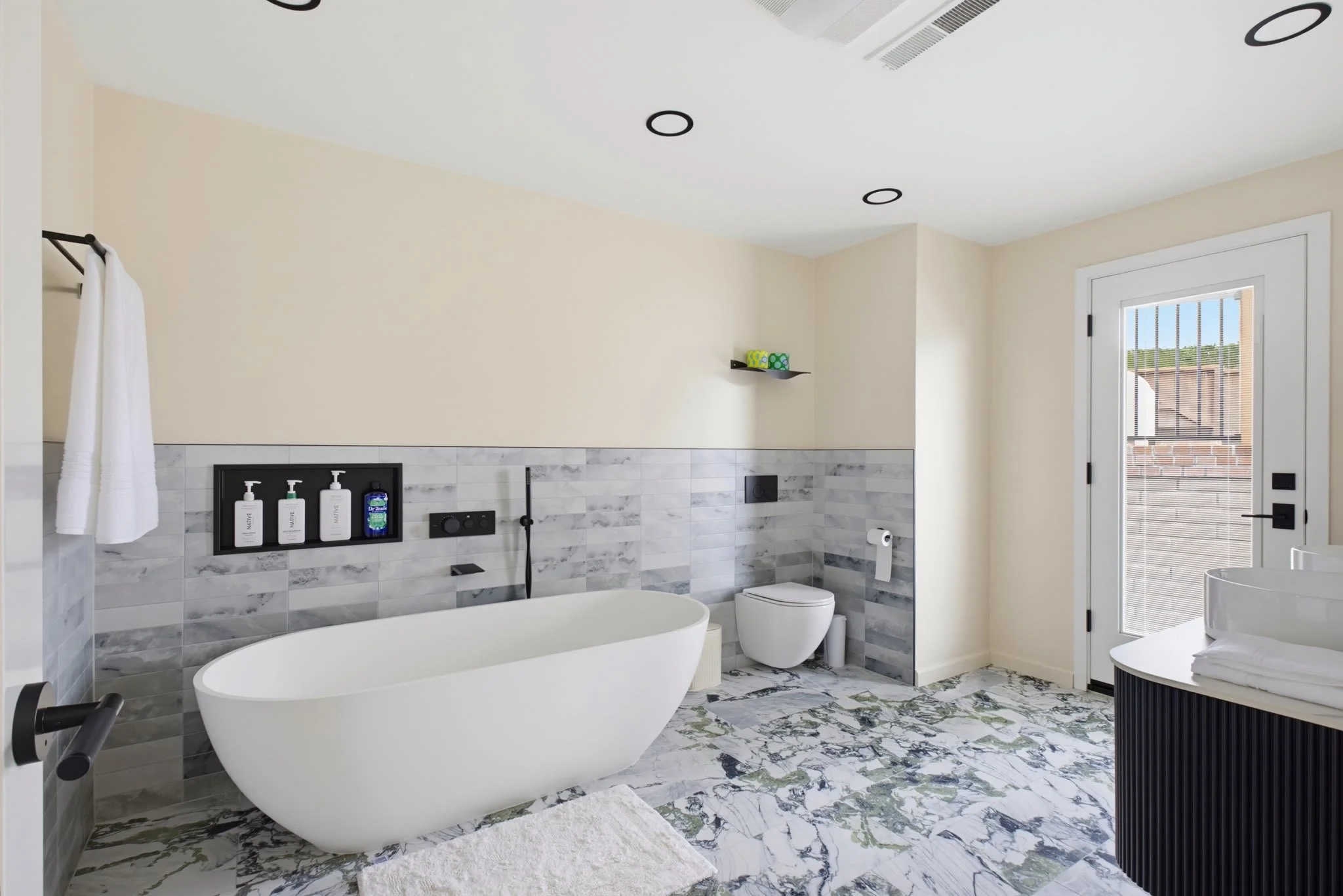 Modern bathroom with white freestanding bathtub, marble tile floor, and a beige wall with a black shelf holding toiletries. There is a white toilet, a window with blinds, and a circular white side table. The ceiling has black circular recessed lights.