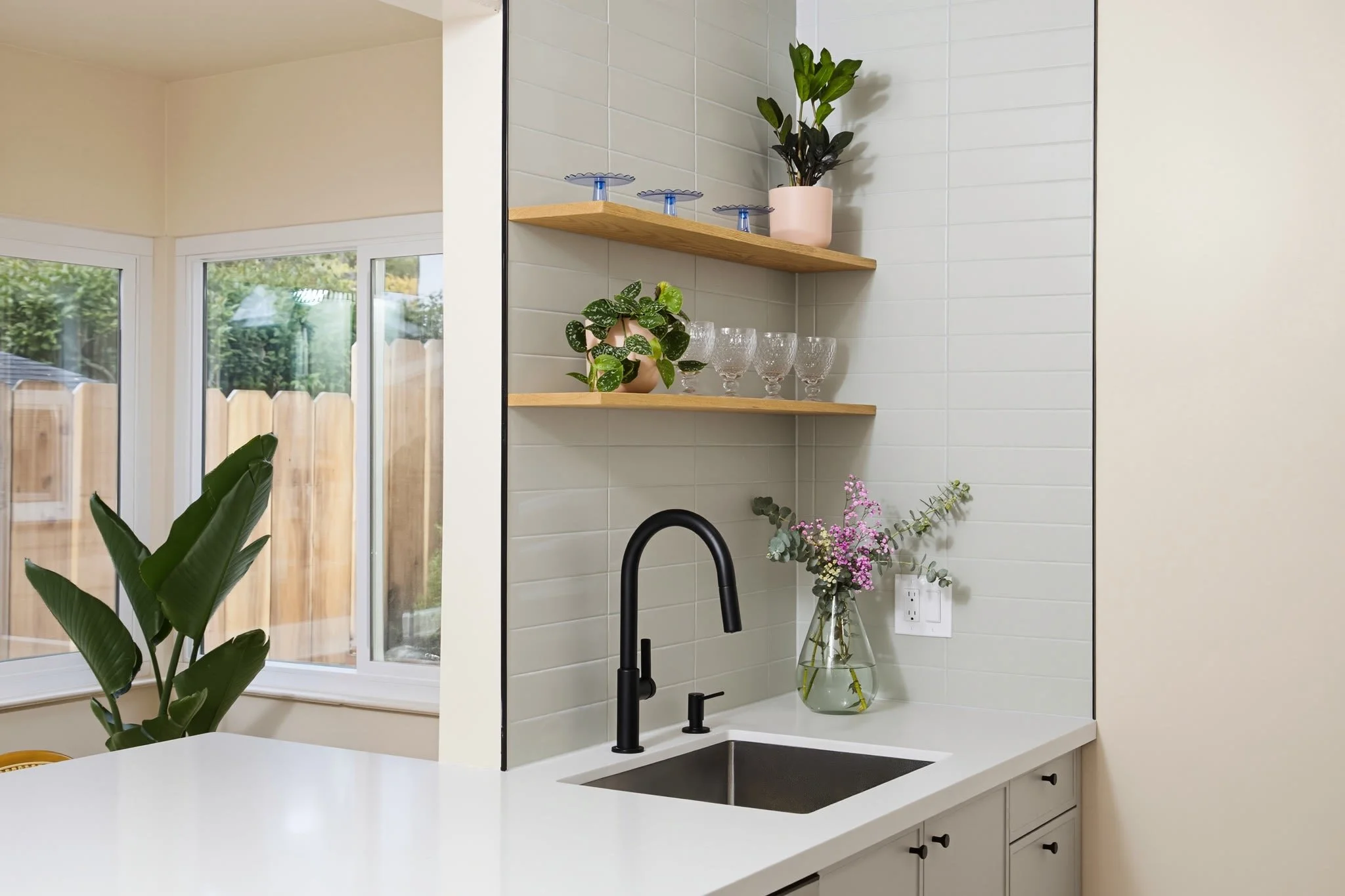 A modern kitchen with white cabinetry, a white countertop, a black faucet, and decorative plants on wooden shelves and a countertop, with a large window showing green trees outside.