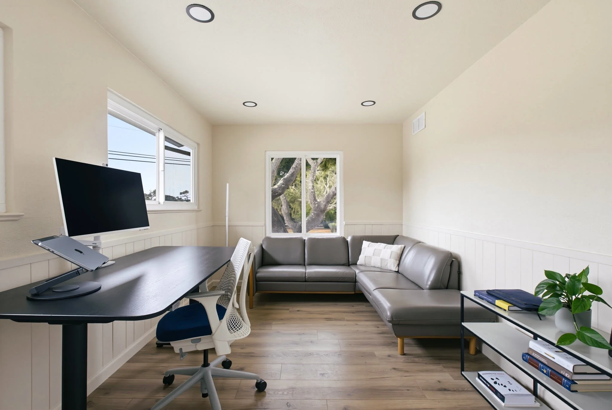 Bright living room with a gray sectional sofa, a window showing trees outside, a dark desk with a monitor and laptop, a white office chair, and a black and white bookshelf with books and decorative items.