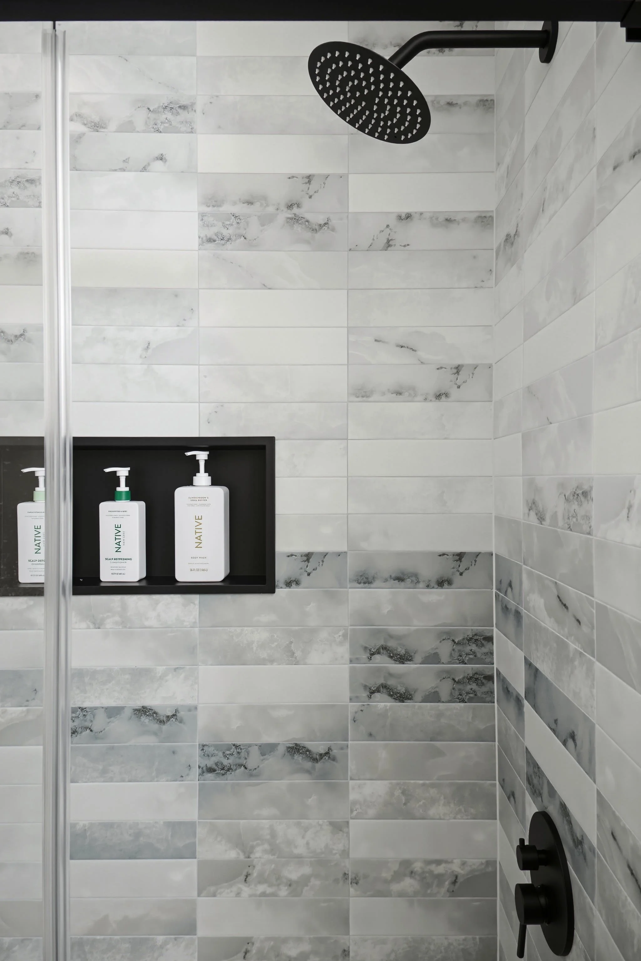 Shower with black rain shower head, gray marble wall tiles, and a side shelf holding three bottles of personal care products.
