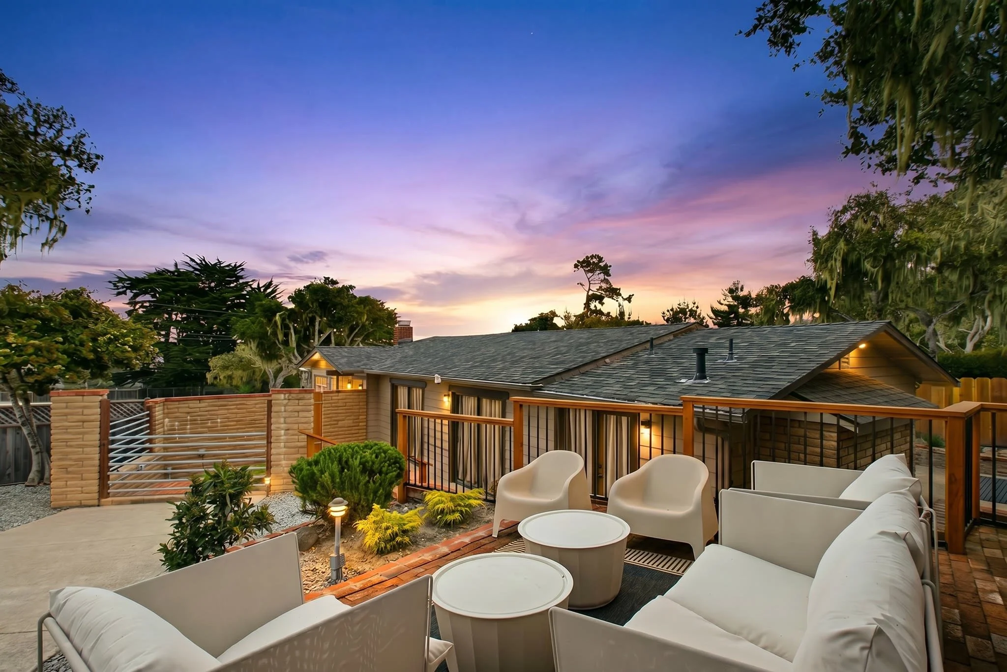 A backyard patio with modern white outdoor chairs and round tables, surrounded by plants, a brick wall, and a wooden railing. The sky is at sunset with shades of purple, pink, and blue, and neighboring houses are partially visible.