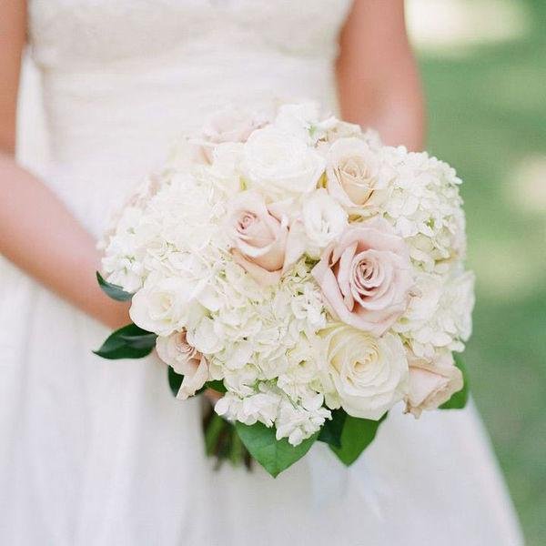 A bride holding a bouquet of white and pale pink roses and hydrangeas, wearing a wedding dress.