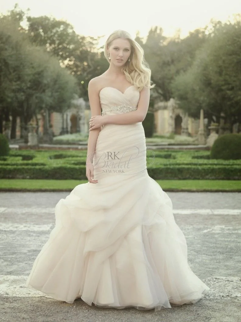 A woman in a strapless white wedding gown standing outdoors on a gravel path with trees and a historic fountain in the background.