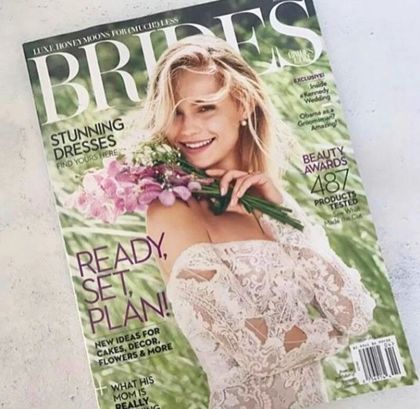 Magazine cover featuring a woman with blonde hair holding a bouquet of pink flowers, smiling and wearing a lace dress, with magazine headlines about dresses, beauty awards, and plans.