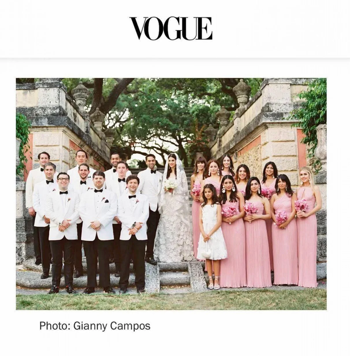 A large wedding party group photo outdoors with stone structure and trees in the background. The bride and groom are standing in the center, surrounded by groomsmen in white tuxedos and women in pink dresses holding pink bouquets.