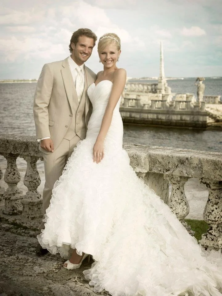A newlywed couple dressed in wedding attire, standing near a stone railing by the water with a pier and cloudy sky in the background.