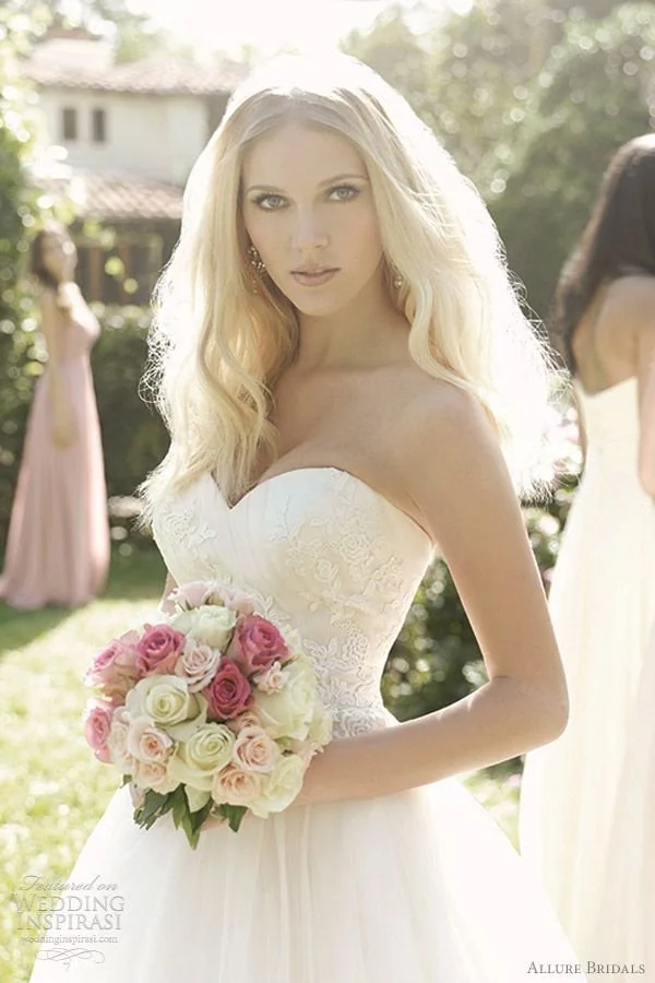 A bride in a white strapless wedding dress holding a bouquet of pink and white roses, standing outdoors sunny day with greenery and other women in dresses in the background.