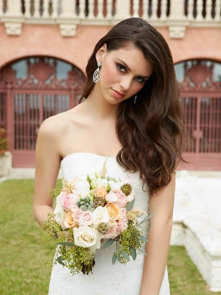 A young woman in a white strapless wedding dress holding a bouquet of pink, white, and peach flowers with greenery, standing outdoors in front of a pink building with arched windows and a green lawn.