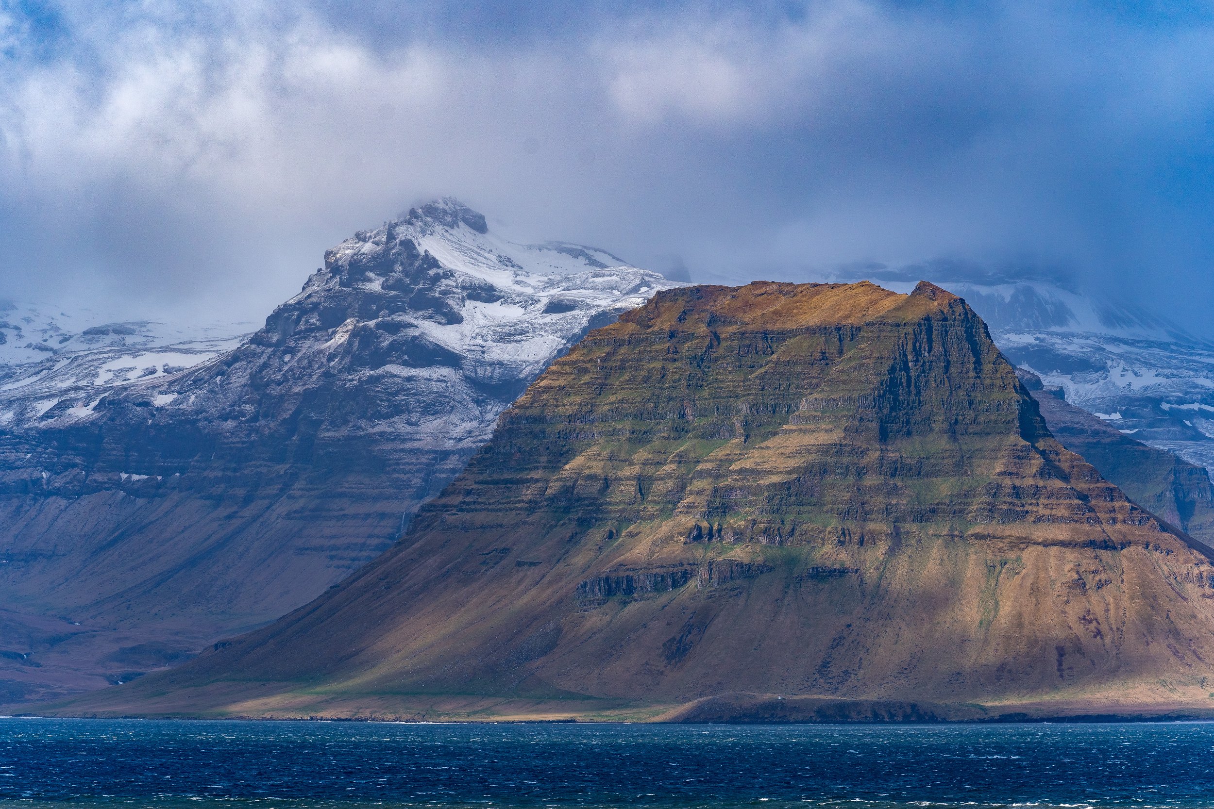 A mountain landscape with snow-capped peaks, rocky slopes, and a body of water in the foreground.