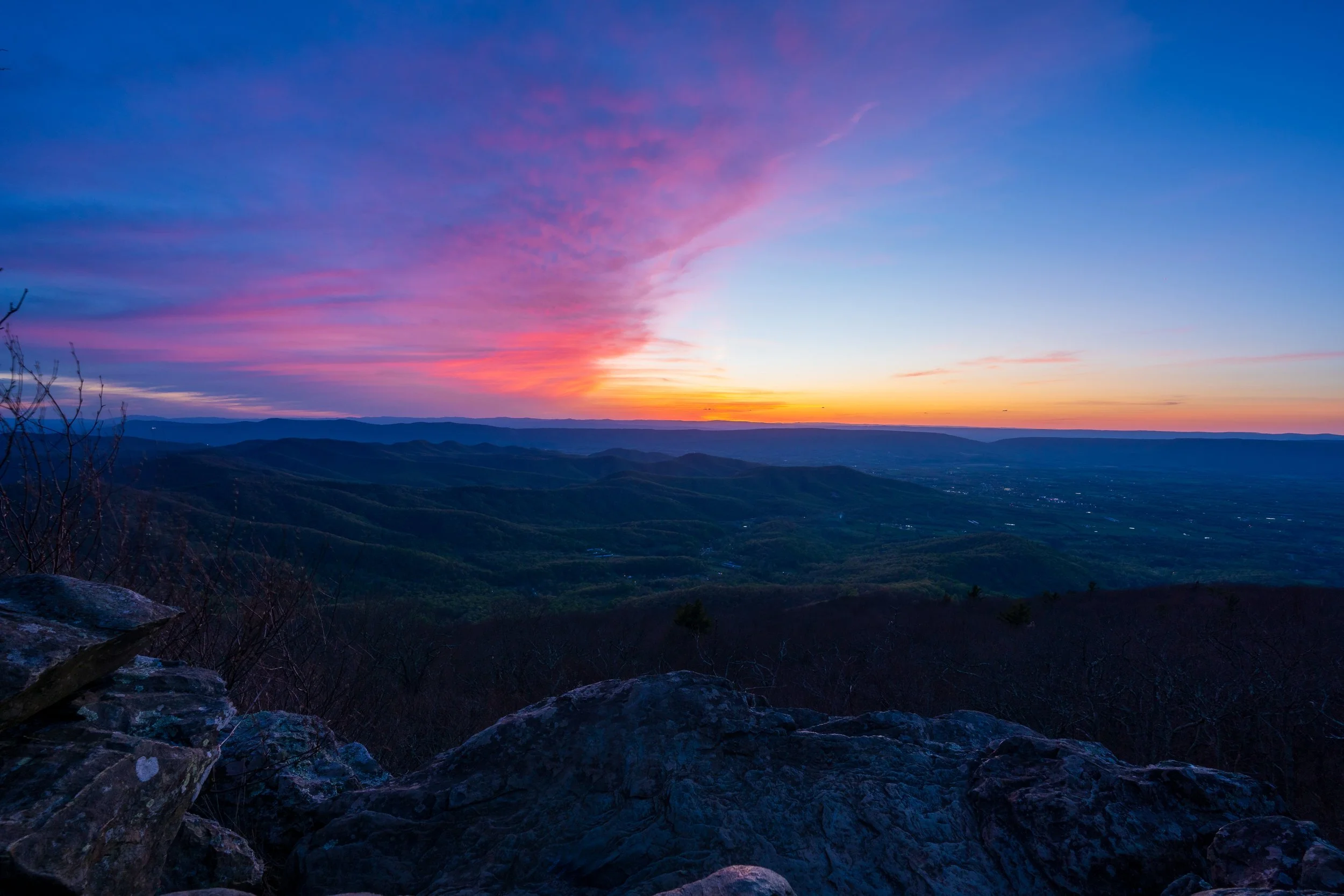 A scenic view of a mountain landscape during sunset with colorful pink, purple, and orange clouds in the sky over distant rolling hills.
