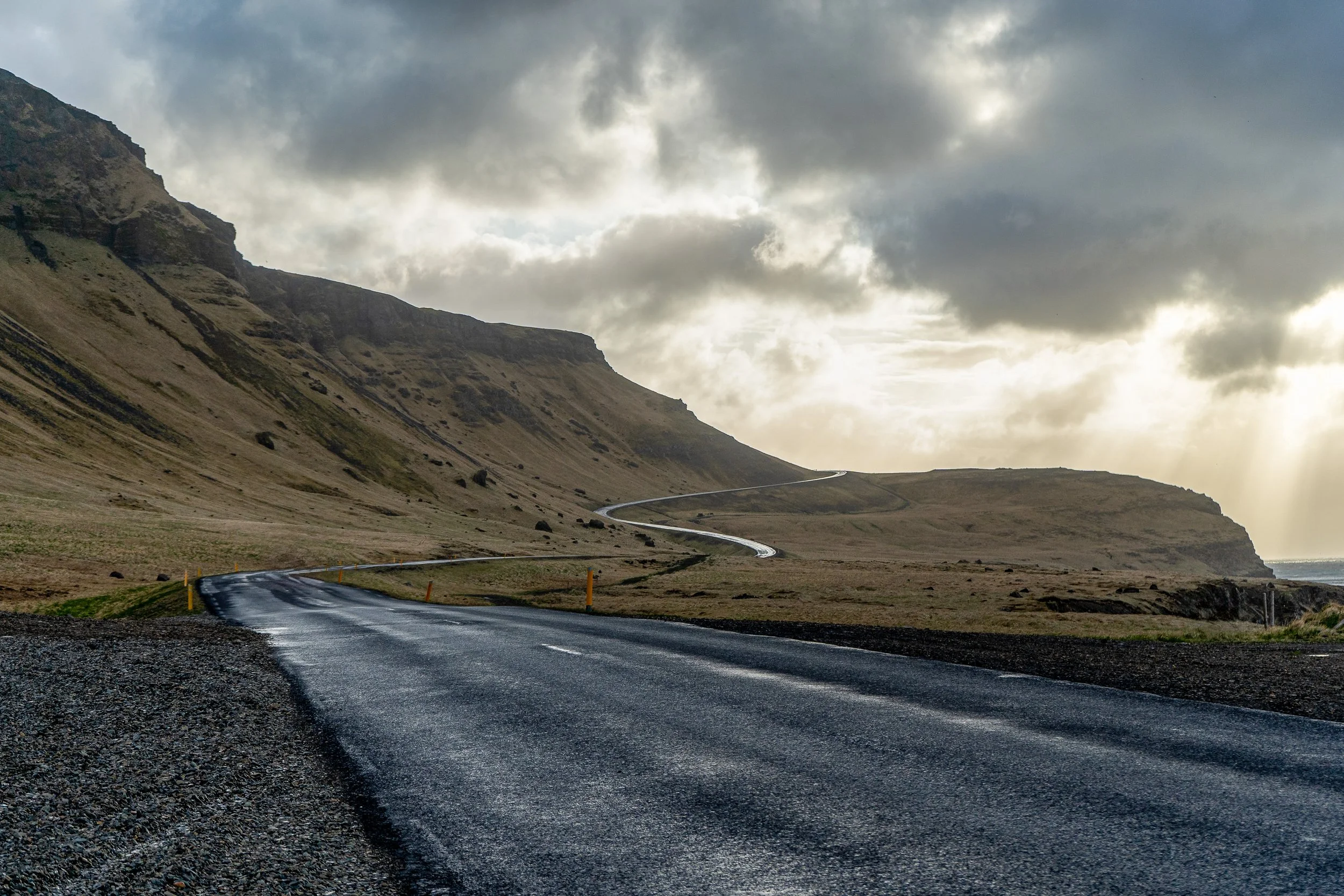 A winding road running through a mountainous landscape under cloudy skies, with patches of sunlight breaking through the clouds.