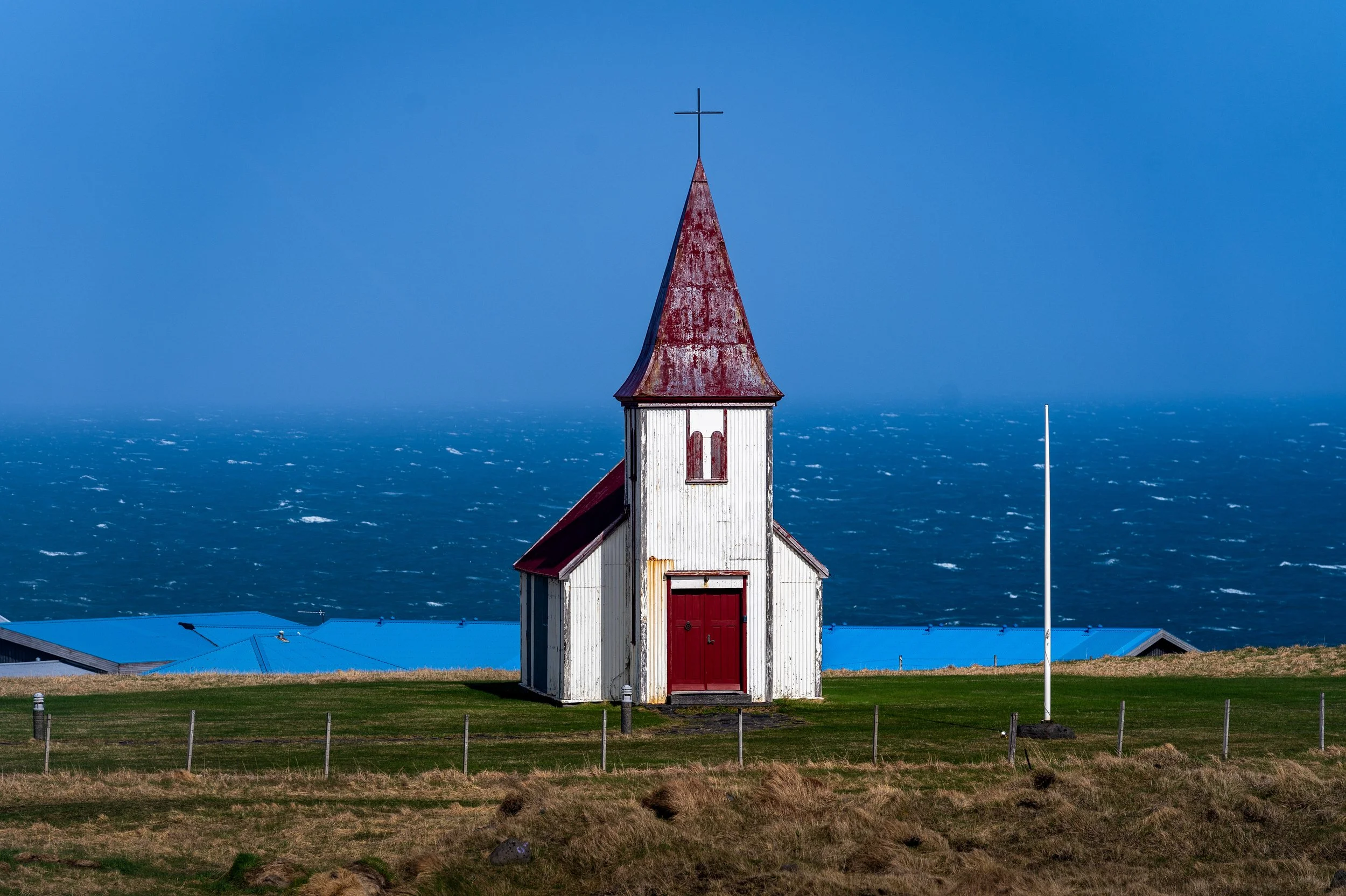 A small white church with a red door and red roof, situated on a grassy hill overlooking the ocean on a windy day.