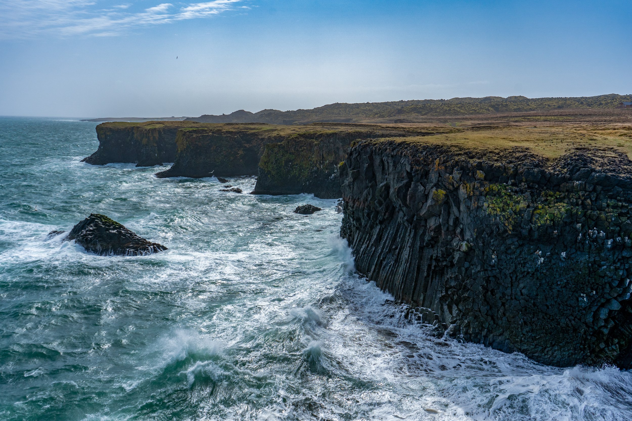 Cliffside coastal landscape with tall, rugged cliffs overlooking the ocean, waves crashing against the rocks below, and a partly cloudy sky.