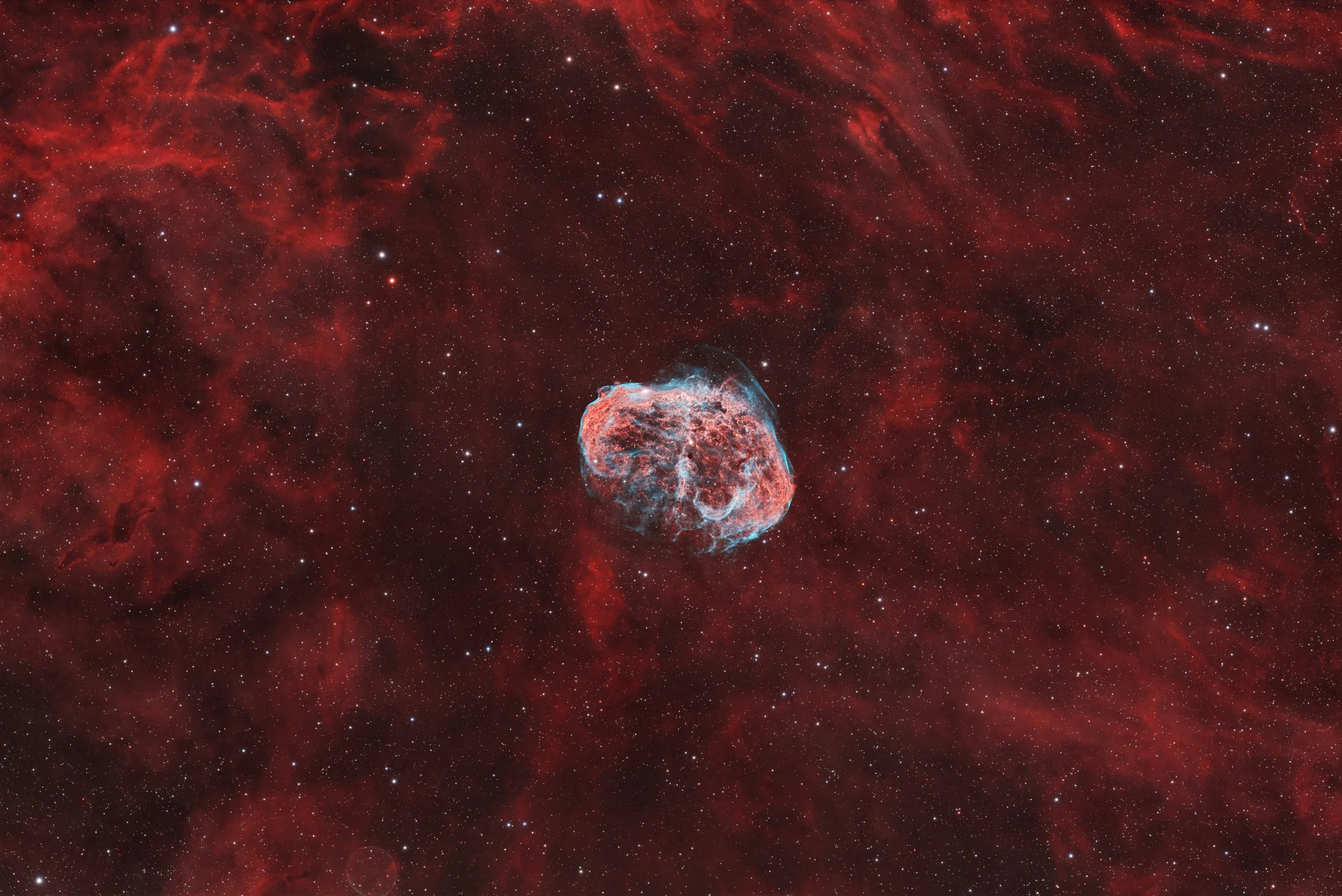 A photograph of a nebula in space with a small, bright, and colorful supernova remnant at the center, surrounded by a vast field of stars and reddish cosmic dust.