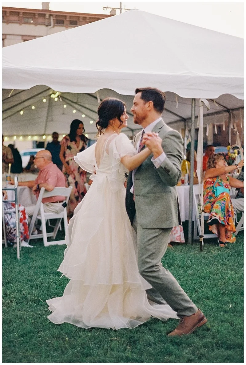 A bride and groom dancing at an outdoor wedding reception under a white tent with guests seated at tables.