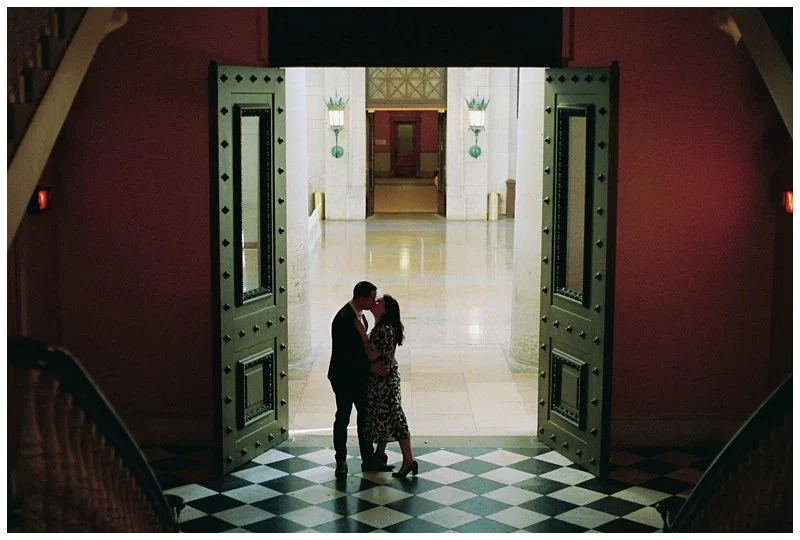 A couple standing close together in an embrace in front of large open doors leading to a grand hallway. The hallway has white marble floors, tall white columns, and decorative wall-mounted lamps, with a set of double doors in the background.