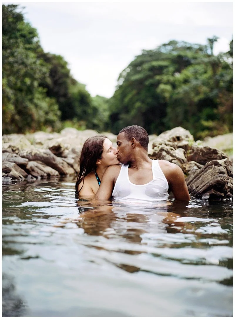 A couple kissing in a river with rocks and green trees in the background.