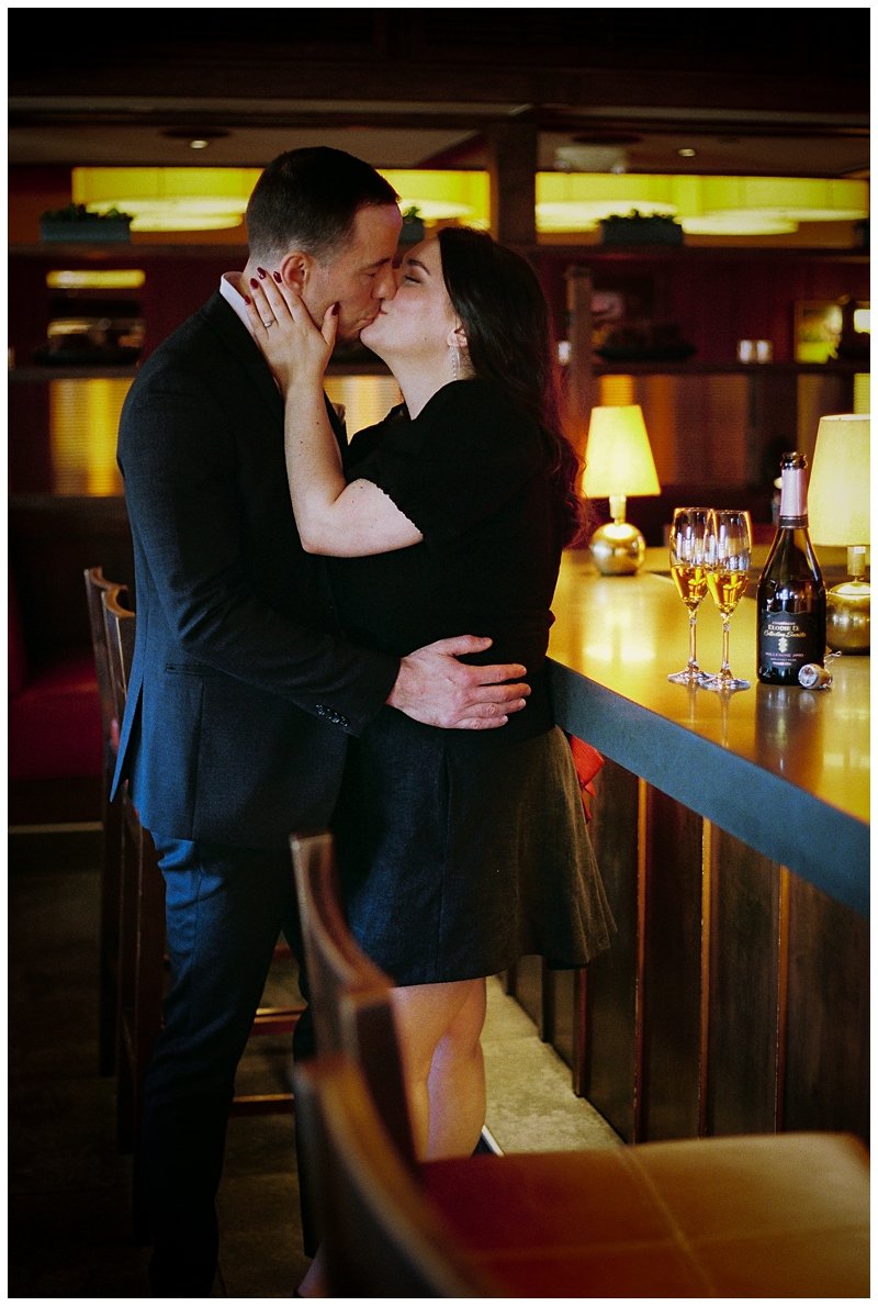A couple kissing in a warmly lit bar with glasses of champagne and a bottle on the counter.