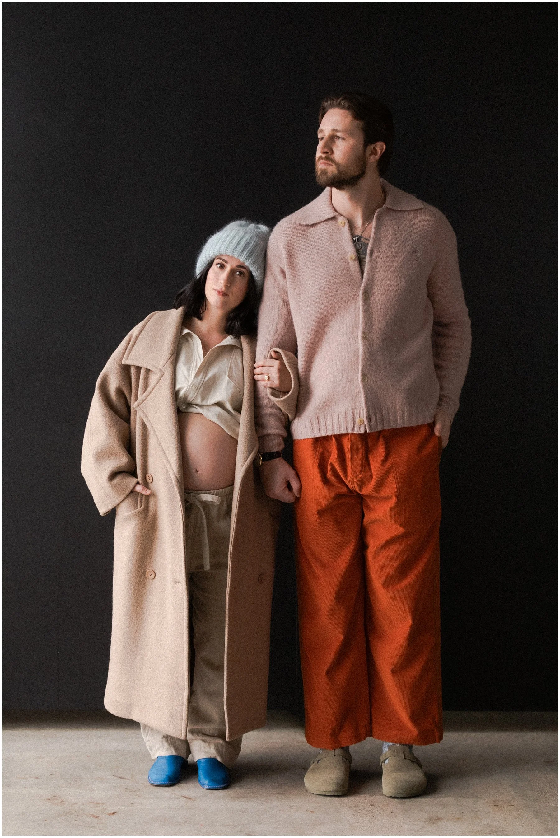 A pregnant woman and a man standing against a dark background. The woman is wearing a long beige coat, a beige shirt tied at her waist, cream-colored pants, blue shoes, and a blue knit hat. The man is wearing a pink cardigan, orange pants, and beige 