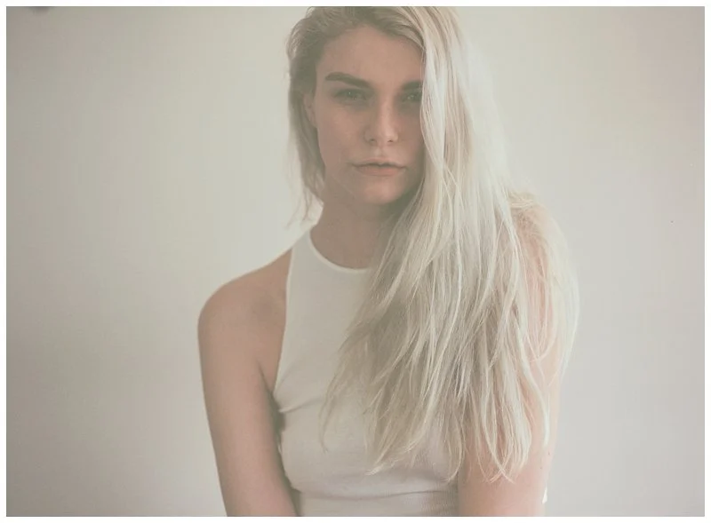 A young woman with long, blonde hair is wearing a white sleeveless top, standing against a plain, light-colored background.