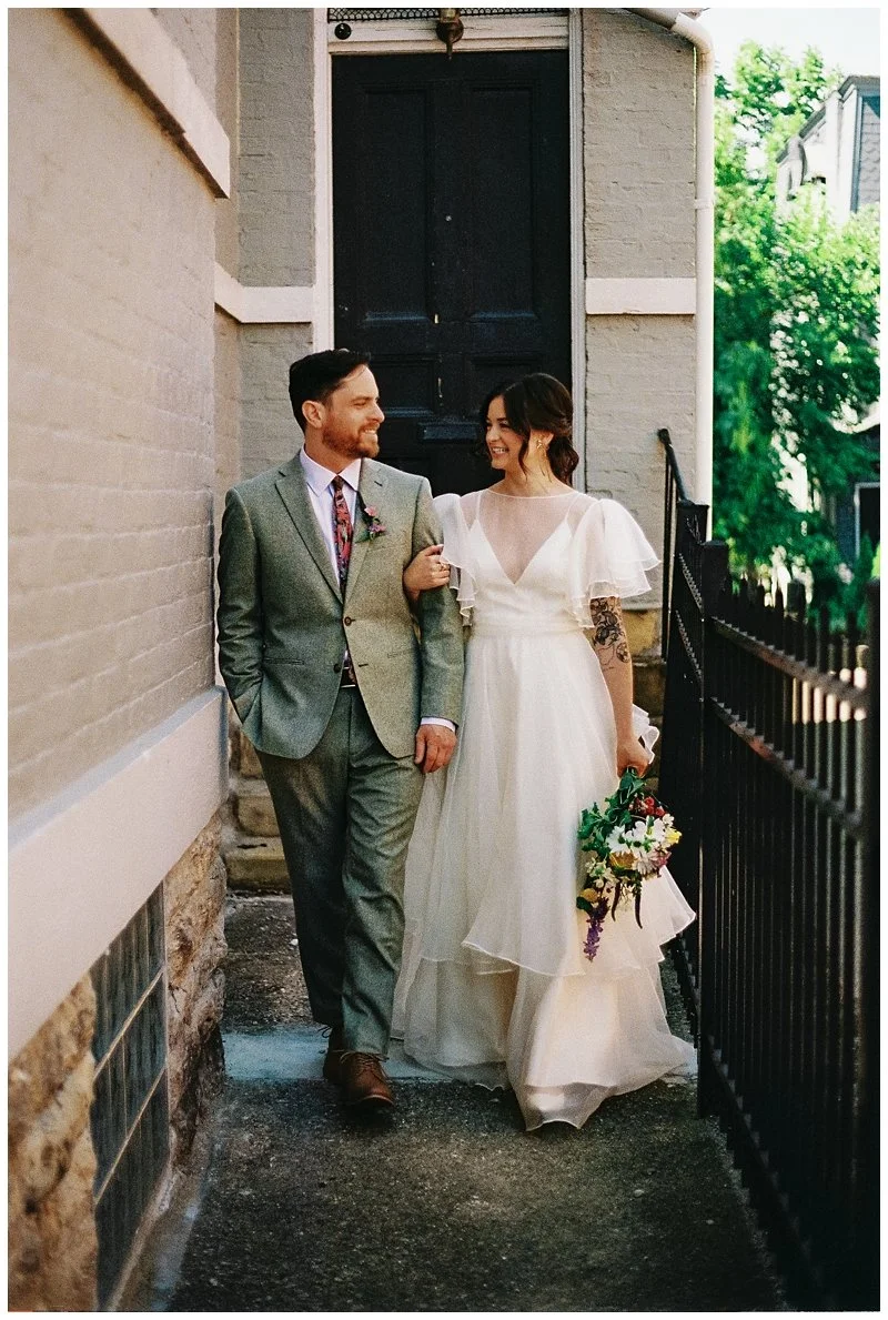 A bride and groom walking arm-in-arm outdoors near a black door and black wrought iron fence, smiling at each other. The bride is wearing a white wedding dress and holding a bouquet, and the groom is dressed in a gray suit with a colorful tie. Film P