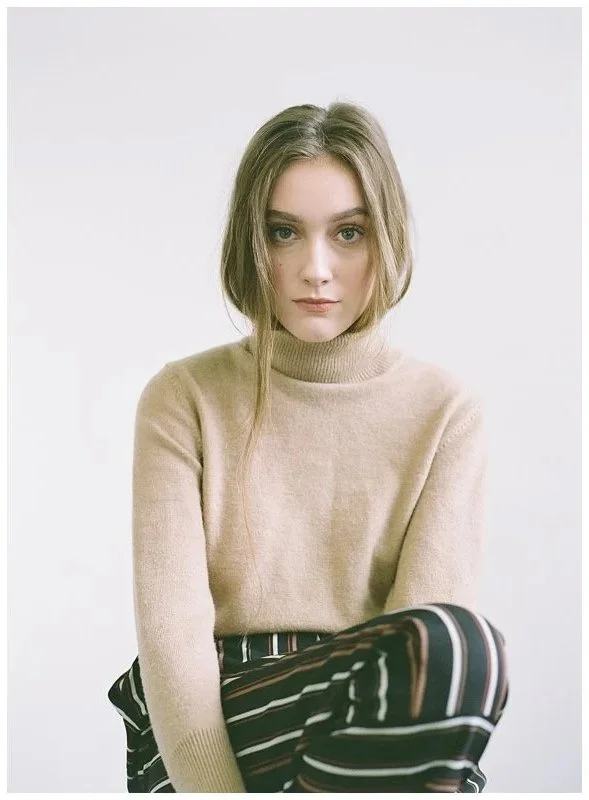 A young woman with shoulder-length light brown hair and blue eyes sitting against a plain white background. She is wearing a beige turtleneck sweater and striped trousers with black, white, and brown lines.