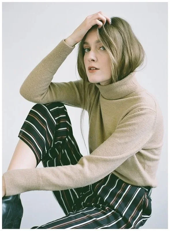A young woman with shoulder-length light brown hair, wearing a beige turtleneck sweater and black pants with colorful vertical stripes, sitting against a plain white background, resting her head on her hand.