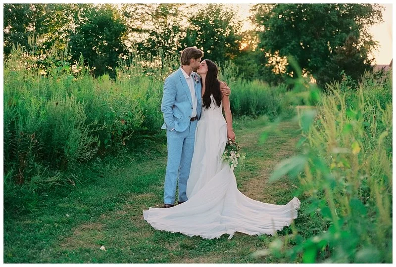 A couple dressed in wedding attire sharing a kiss outdoors in a green field with trees and sunlight in the background.